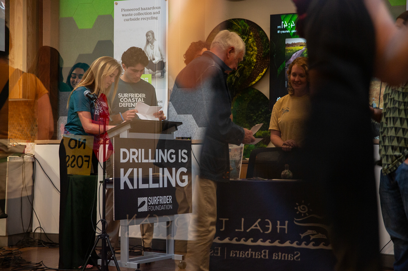 The reflection of people as they stand and mingle during a town hall. Two of those people stand behind a podium with a sign that reads, "drilling is killing" as they look at various sheets of paper.