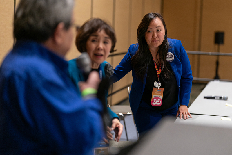 Three people stand behind a set of tables at an event. The photo focuses on an individual wearing a navy pant suit as they reach for the chair behind them. The person next to them looks towards another person holding a microphone.