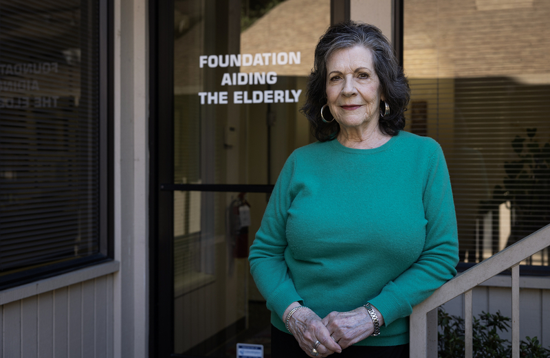 An older adult wearing a green sweater stands outside a glass door with the words “Foundation Aiding the Elderly” printed on it, hands clasped and looking toward the camera.
