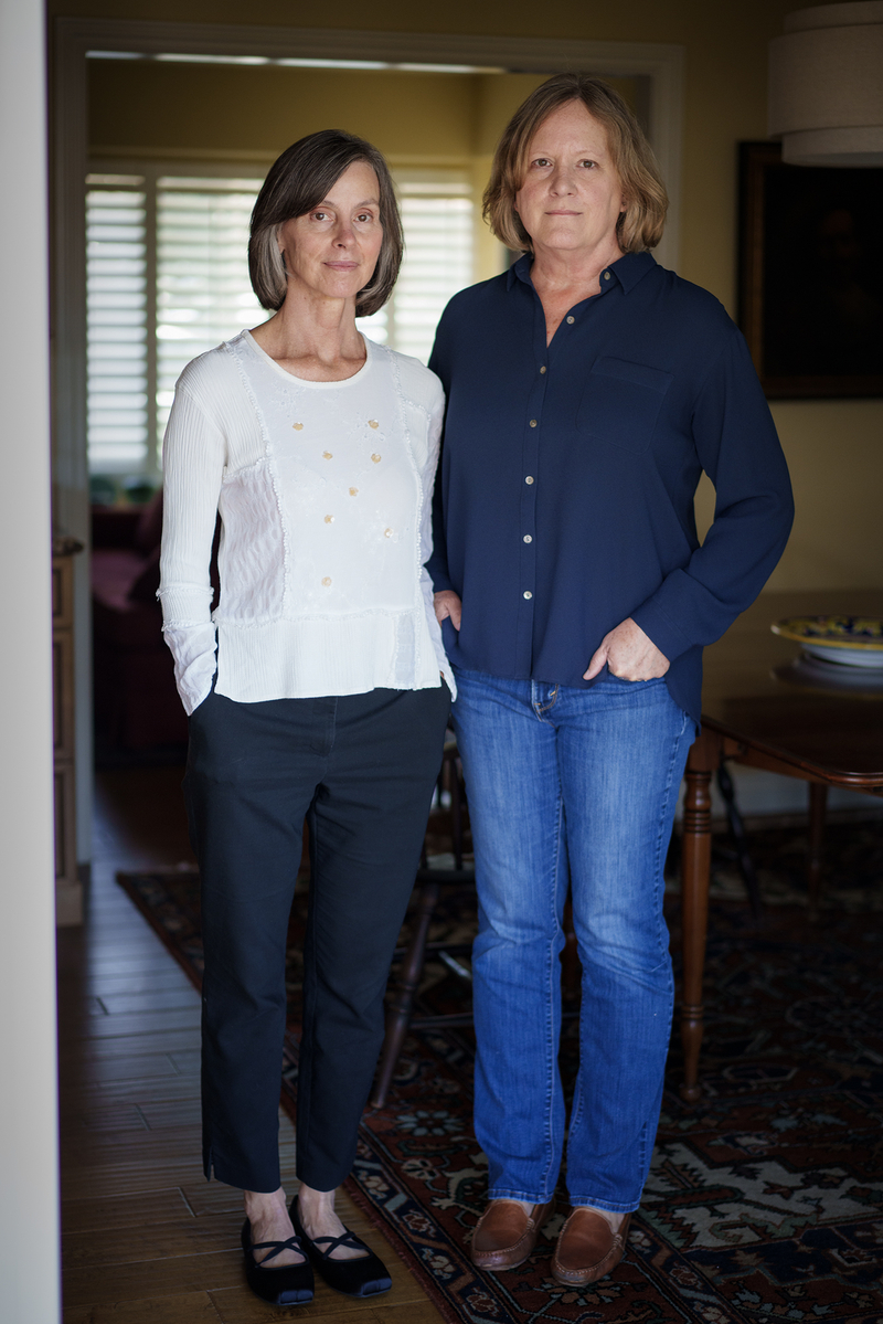 Two people stand side by side indoors, facing the camera. One wears a light long-sleeve top and dark pants, and the other wears a dark button-up shirt and jeans, with hands in their pockets. A doorway with window blinds and a softly lit room appears behind them.