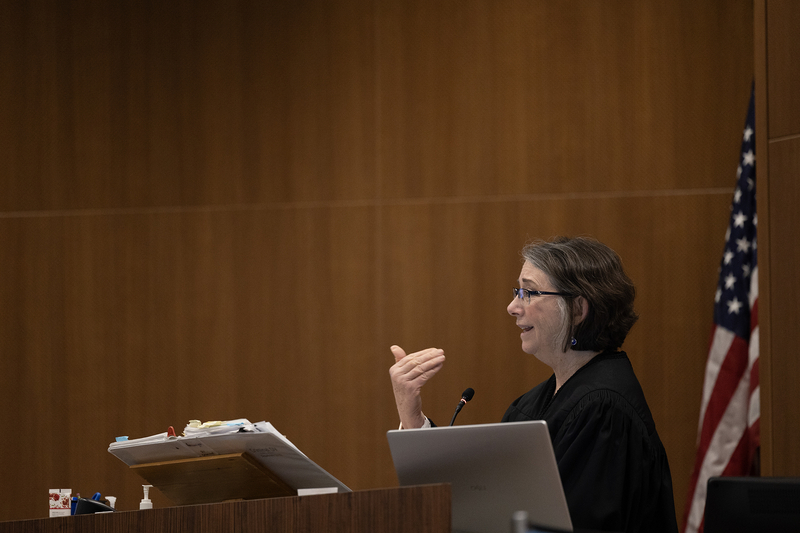 A judge in a black robe speaks from the bench inside a courtroom, gesturing with one hand while addressing the room. Papers, a laptop, and a microphone sit on the desk, and a U.S. flag stands in the background against wood-paneled walls.