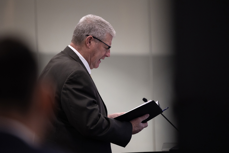 A person in a suit stands at a microphone reading from a notebook during a court proceeding, viewed from the side. The background is softly lit and out of focus.