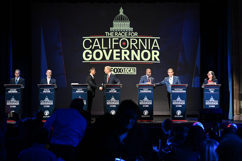 Several candidates stand at podiums on a stage during a televised debate titled “The Race for California Governor,” with a large screen and the Fox Local logo behind them. Some candidates shake hands while others stand at their lecterns as audience members sit in the foreground.