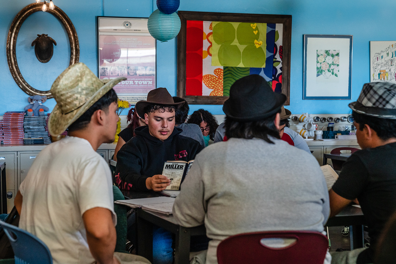 A student wearing a brown cowboy hat reads a book while sitting next to other students inside a classroom.