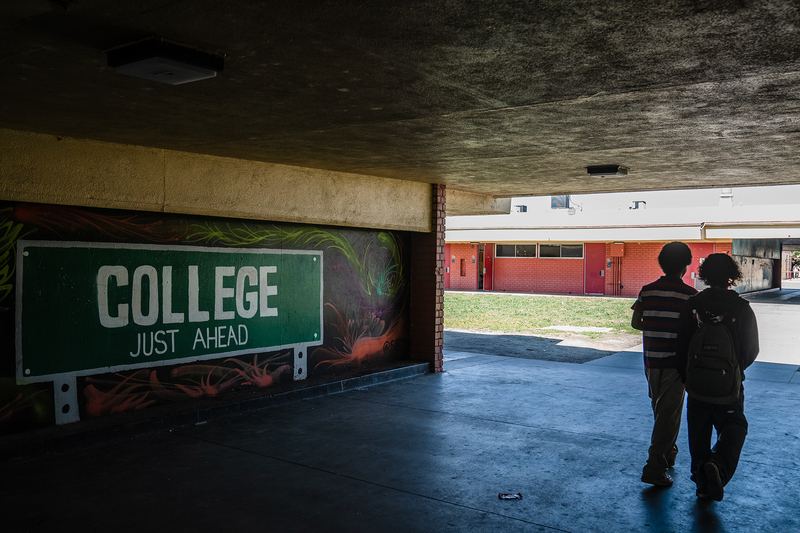 The shadow of two students walking pass a mural of a street sign named "College just ahead."