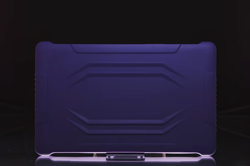 The back of a rugged tablet is shown on a small stand, its matte surface featuring angular, molded ridges and a protective design. Purple lighting outlines the edges and base, casting a soft glow and reflection on the glossy surface beneath, emphasizing a durable, industrial look.