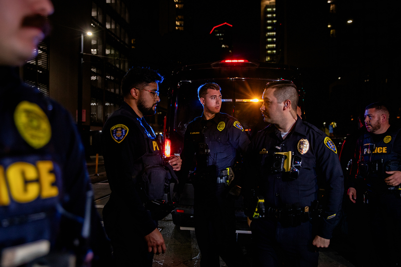 Several police officers face each other as they stand in front of a moving police van in the middle of a street during an evening.