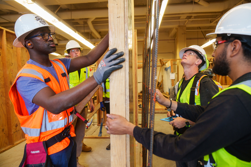A group of students wearing safety vests and hard hats stands around, working on a section of wall construction and rebar near a window in a classroom.