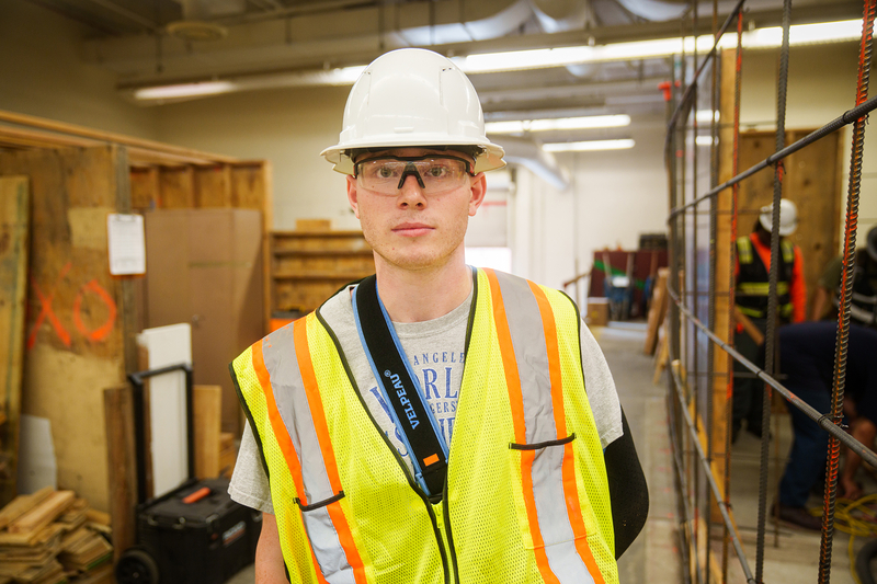 A person wearing a safety vest and a hard hat stands next to a section of rebar inside a classroom with students working around them.