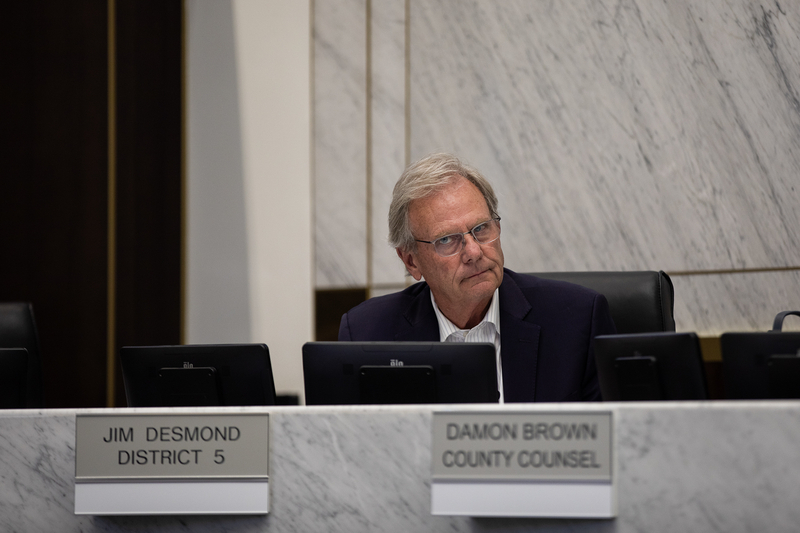 San Diego County Supervisor Jim Desmond sits behind a marble dais during a meeting, looking to his left. Several monitors and nameplates are positioned in front of him, with a marble wall in the background.