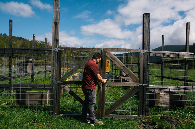 A person, wearing a red shirt and gray pants, locks the wooden gate of a garden at a school overlooking mountains.