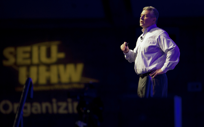 A person stands on a stage under dramatic lighting, speaking into a headset microphone while holding a remote. They face the audience with one hand on their hip, mid-presentation. In the background, a large screen displays blurred text that reads “SEIU UHW,” suggesting a labor or union event.