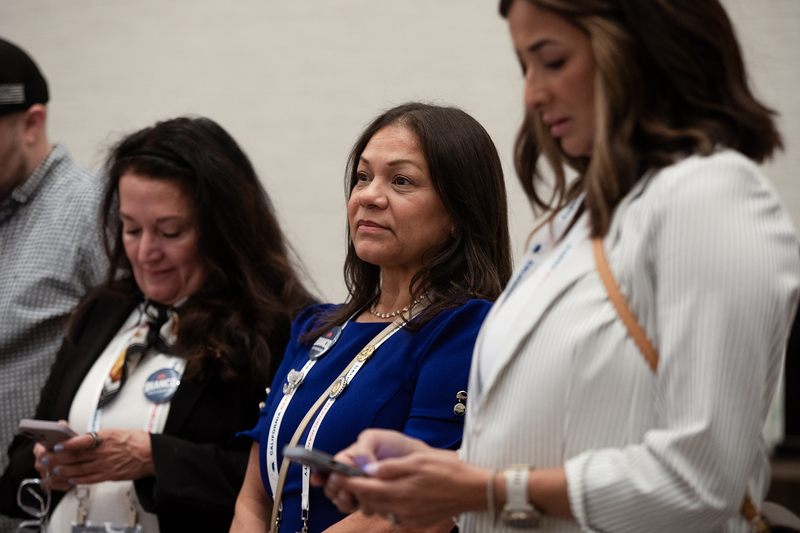 Three people stand side by side indoors, wearing conference badges on lanyards. The person in the center, dressed in a blue top, looks ahead with a composed expression, while the two on either side glance down at their phones. The background is plain and softly lit, keeping the focus on the group.