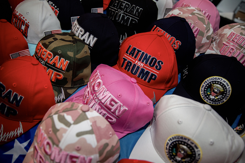 A collection of colorful baseball caps is arranged closely together on a table, many featuring political slogans and logos. Prominent among them are red, pink, camouflage, and black hats embroidered with phrases like “Latinos for Trump” and “Veteran for Trump,” alongside caps bearing an emblem resembling the presidential seal.
