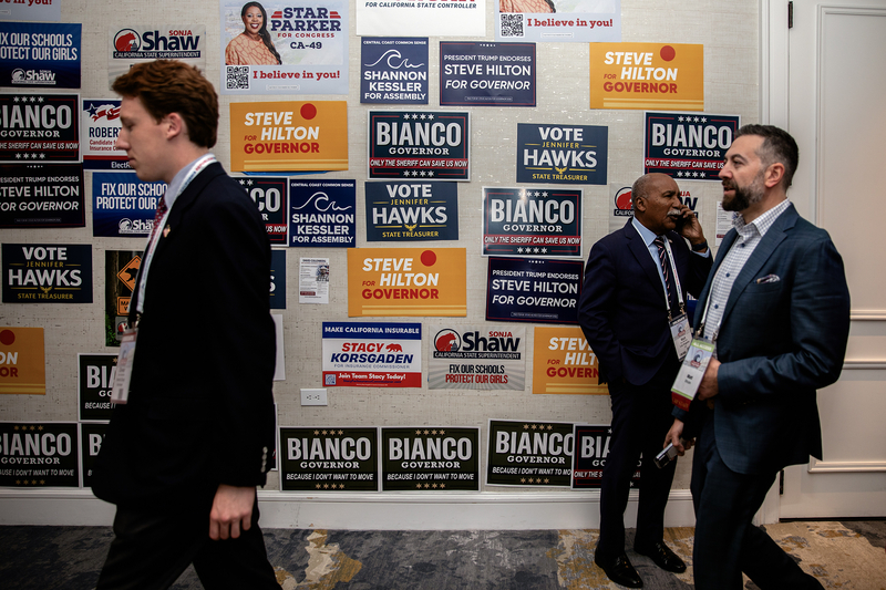 Three people in business attire move through a hallway lined with a wall covered in political campaign signs. One person walks past in the foreground, while two others stand and talk, one holding a phone to their ear. The signs behind them promote various candidates and offices, with bold colors and slogans filling the background.