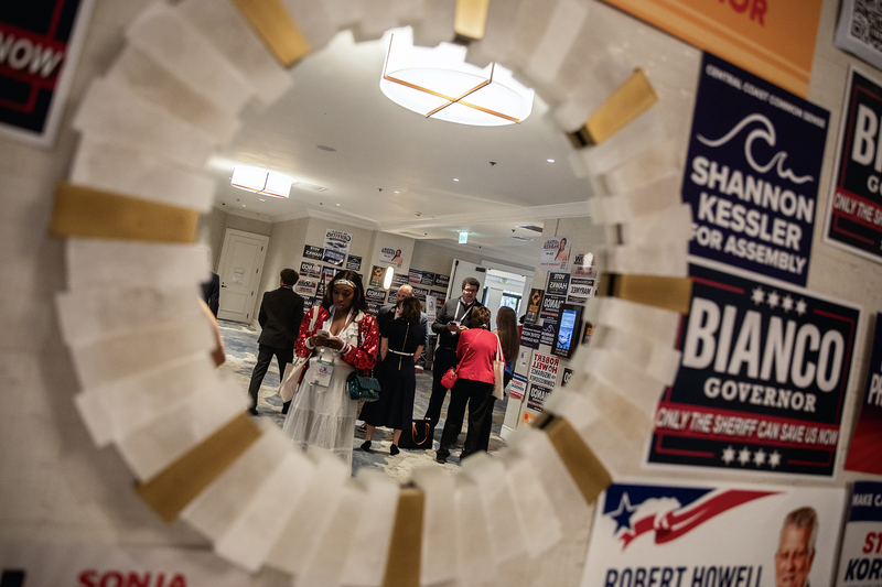 A group of people stands in a hallway at a political event, seen through the circular reflection of a decorative mirror. Several attendees wear conference badges and look down at their phones while others talk nearby. The walls around them are covered with campaign signs for various candidates, creating a busy, layered backdrop.