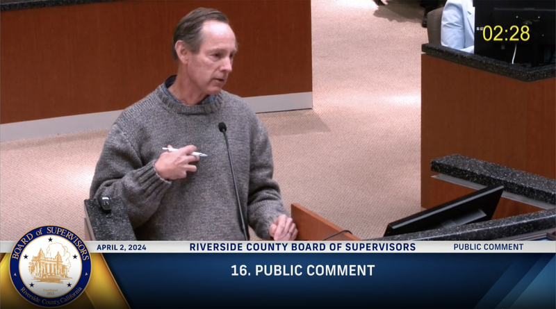 A person stands at a podium during a Riverside County Board of Supervisors meeting, speaking into a microphone while holding a pen. They wear a gray sweater and face slightly to the side. On-screen graphics show “APRIL 2, 2024,” “RIVERSIDE COUNTY BOARD OF SUPERVISORS,” and “16. PUBLIC COMMENT,” with a countdown timer in the upper corner indicating time remaining to speak.
