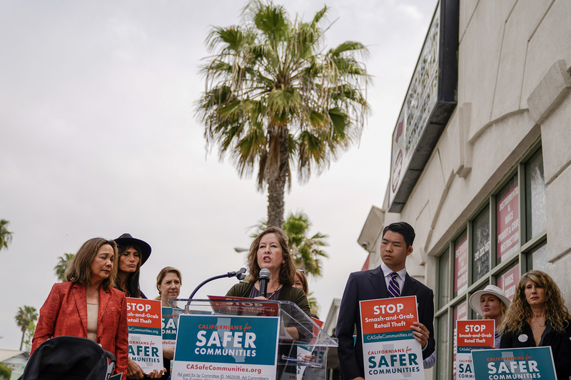 A person speaks at a podium during an outdoor press event, surrounded by a group holding signs that read “Stop Smash-and-Grab Retail Theft” and “Californians for Safer Communities,” with a palm tree and storefront in the background.