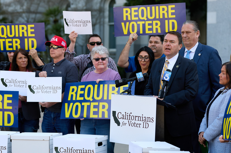 A lawmaker, wearing a black suit with a striped yellow tie, stands behind a lectern while surrounded by people holding blue and yellow signs that say "REQUIRE VOTER ID."