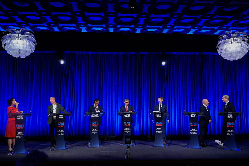 Seven people standing behind podiums up on a stage illuminated by studio light and decorated with a blue curtain.