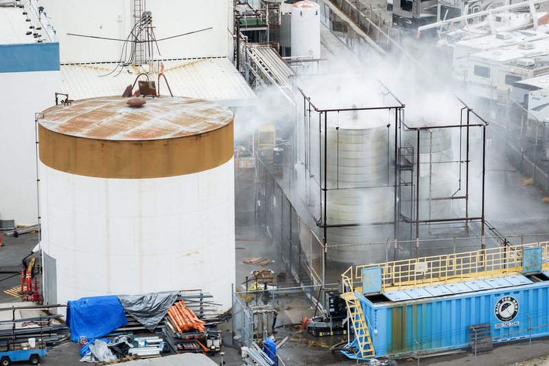 Steam rises from industrial tanks and pipes at a fenced facility site. A large rust-streaked white storage tank stands beside smaller cylindrical tanks releasing vapor, while construction materials, scaffolding and a blue container occupy the foreground. RVs and trailers are parked beyond the facility in the background.