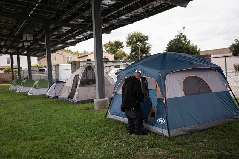 A row of camping tents sits on a grassy field beneath a large solar panel canopy. In the foreground, a person dressed in dark clothing stands at the entrance of a blue and gray tent, partially turned away from the camera. Additional tents stretch into the background alongside a chain-link fence, with houses and recreational vehicles visible beyond the site.