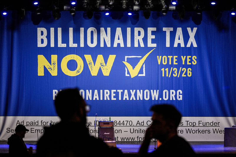 A large stage backdrop reads "Billionaire Tax Now" and "Vote Yes 11/3/26" as attendees gather in silhouette before an event, with musical equipment set up onstage.