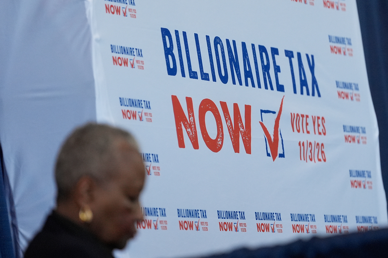 A large white, blue and red sign is displayed at an event. The sign reads " Billionaire Tax Now" and "Vote Yes 11/3/26" next to an image of a check box. A person can be seen walking by in the blurred foreground.