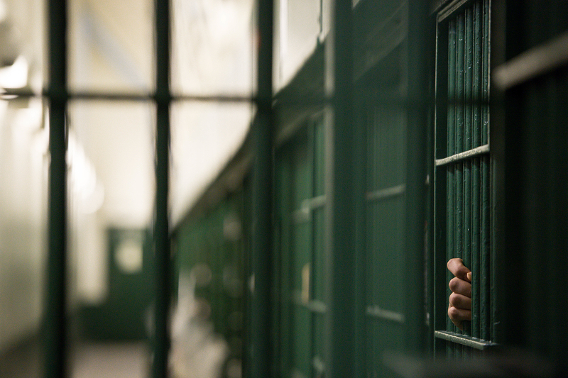 An unidentifiable person's hand grips onto one of the green bars of a jail cell.