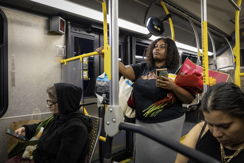A bus rider stands near the rear door holding a bouquet of flowers, shopping bags, a drink and a cellphone while gripping a yellow handrail. Other passengers sit nearby as the evening bus continues along its route.