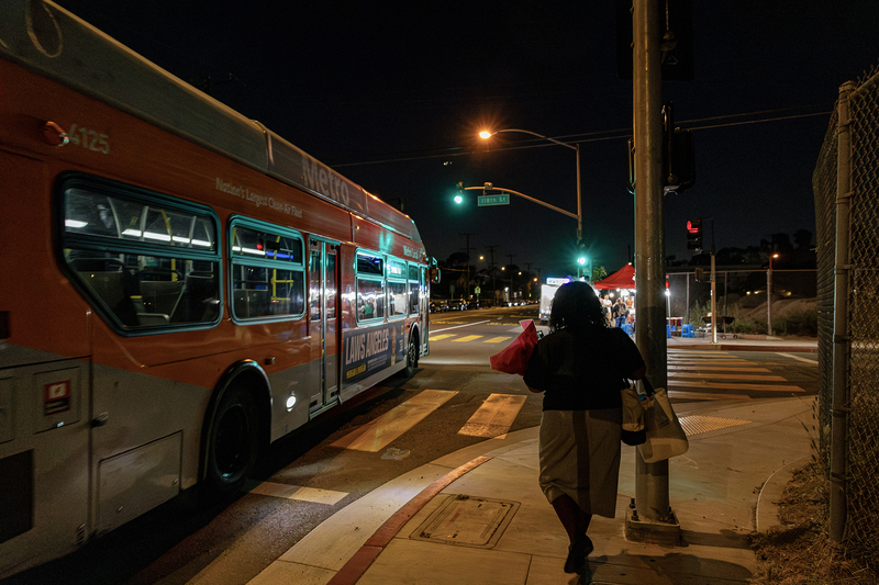 A bus rider carrying shopping bags and a bouquet of flowers walks along a sidewalk at night after stepping off a Metro bus. Streetlights, traffic signals and a brightly lit sidewalk vendor illuminate the intersection ahead.