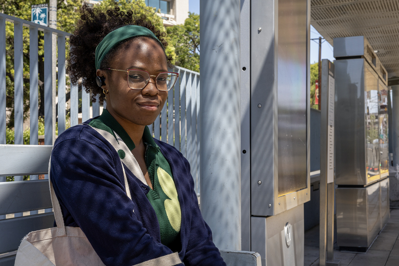 A transit rider wearing glasses, wireless earbuds and a green headband sits on a bench at a Metro rail station platform in daylight, looking toward the camera. Metal information kiosks and the station platform extend into the background.