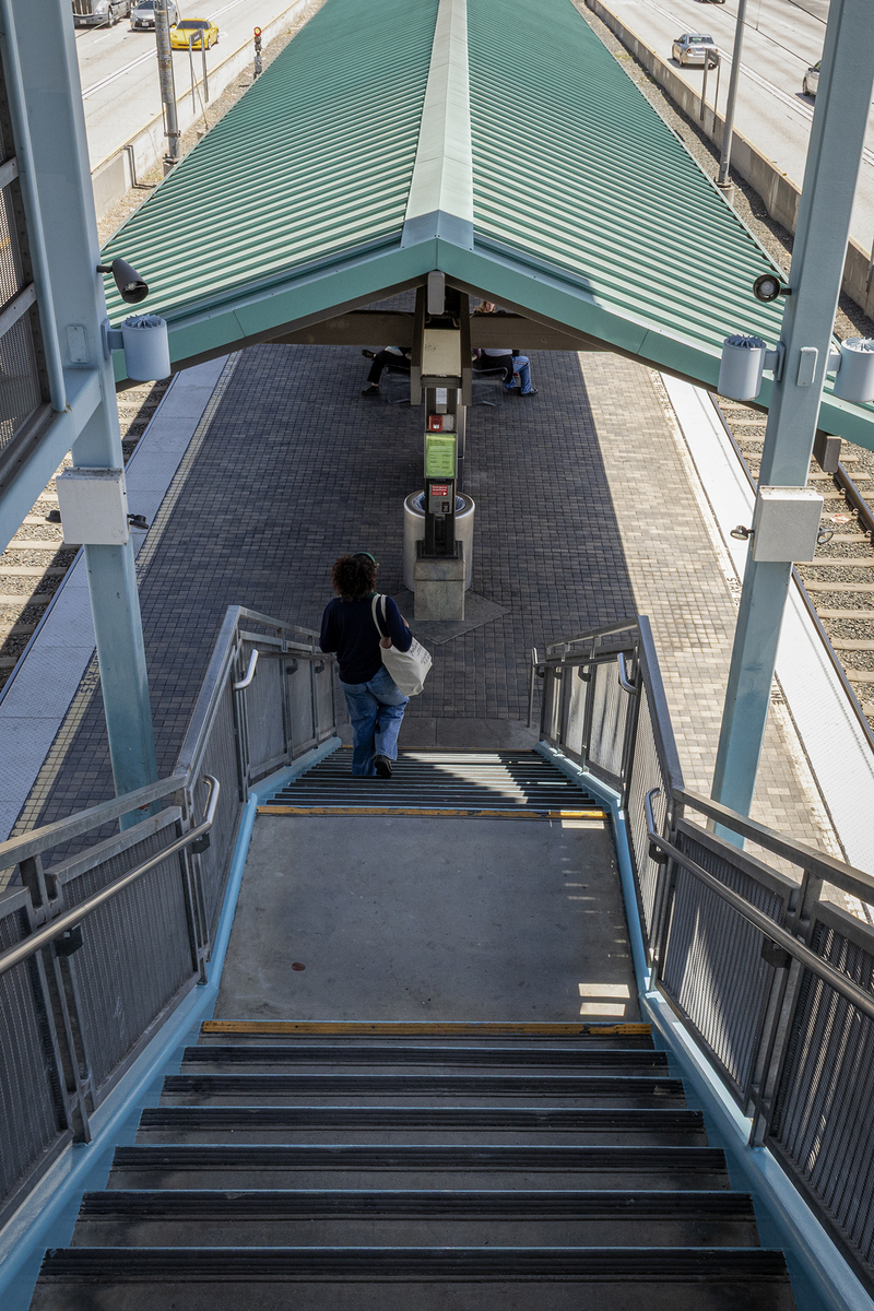 A transit rider carrying a canvas tote bag walks down a staircase from an elevated pedestrian bridge to a Metro rail platform below. The platform is covered by a green metal canopy, with train tracks on either side and a freeway running parallel in the background.
