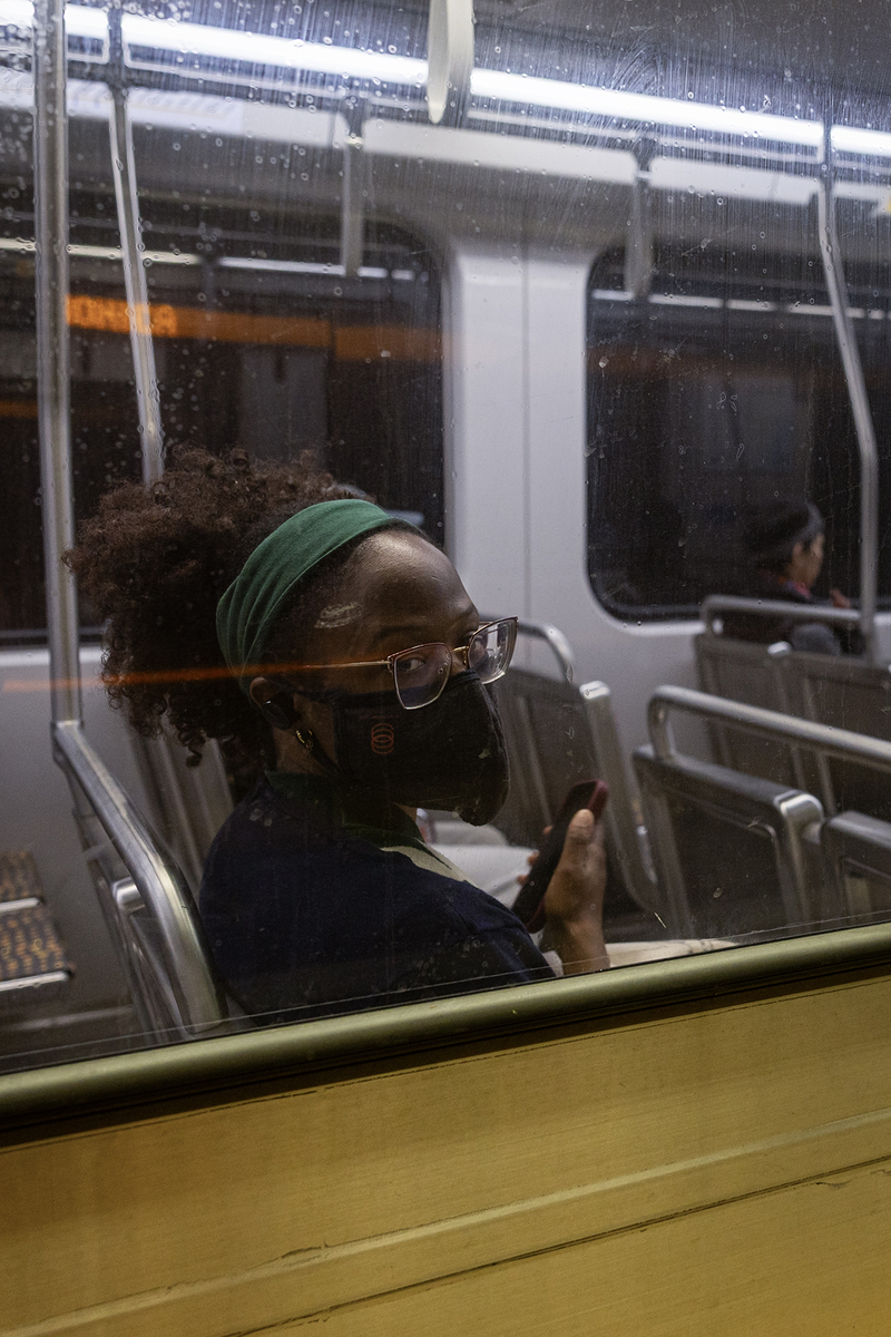 Viewed through the window of a Metro train, a transit rider wearing glasses, a face mask and a green headband sits holding a cellphone and looks toward the camera. Reflections and smudges on the window partially obscure the view, while the mostly empty train car extends into the background.