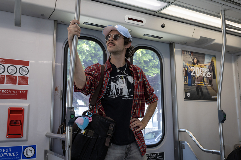 A transit rider wearing sunglasses, a light blue baseball cap and a red plaid shirt stands inside a Metro rail car, holding a vertical pole with one hand and a shoulder bag with the other. Sunlight streams through the train windows behind them, and Metro safety signs and an advertisement line the walls.