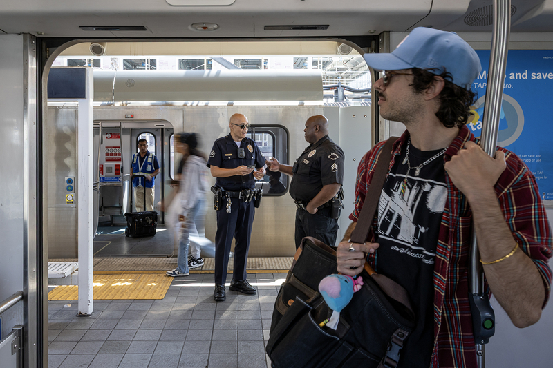 Viewed from inside a Metro rail car, a transit rider wearing a light blue baseball cap, sunglasses and a red plaid shirt stands by the open doorway holding a vertical pole. On the platform outside, two Metro Transit Security officers talk while passengers board and exit another train.