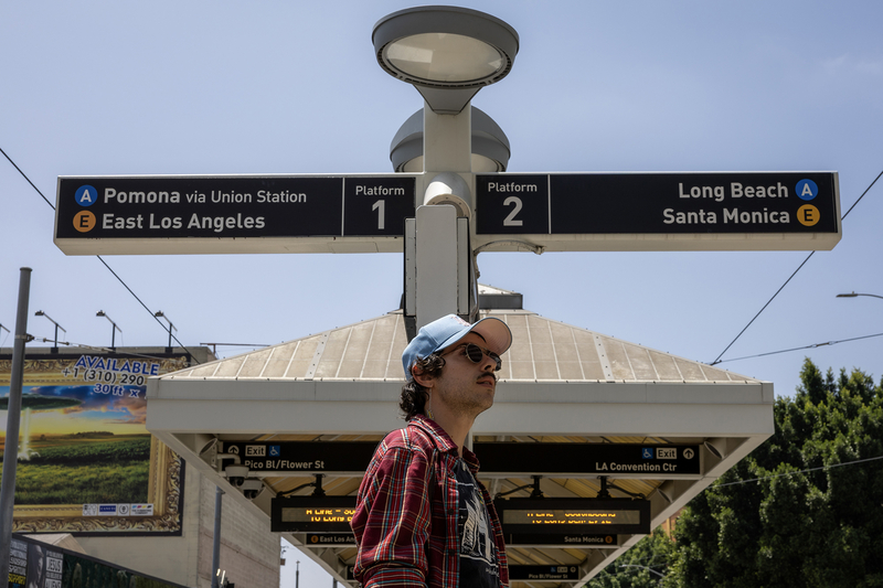 A transit rider wearing sunglasses, a light blue baseball cap and a red plaid shirt stands beneath overhead signs at a Metro rail station showing directions for the A and E lines. The station platforms and canopy extend behind them under a clear blue sky.