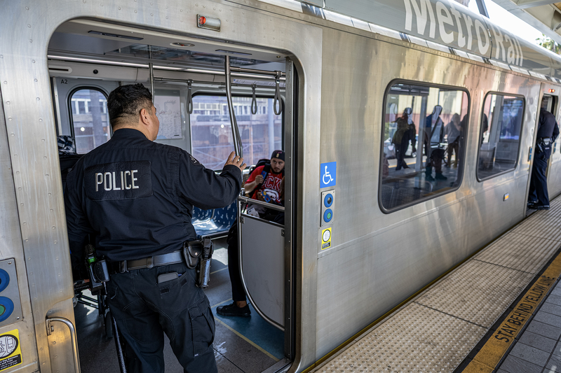 A Metro Transit Police officer stands at the open doorway of a Metro Rail train, speaking with a passenger seated inside. Another officer is visible farther down the platform as passengers and reflections appear in the train windows.
