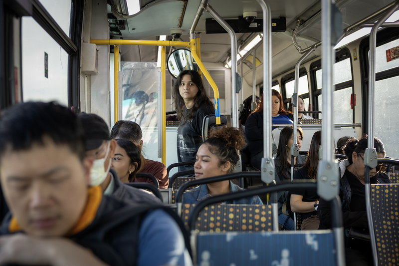 Passengers ride aboard a Metro bus, with several seated while one rider stands near the rear door looking toward the front of the bus. Sunlight streams through the windows, illuminating the crowded interior and rows of seats.