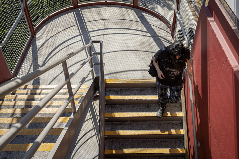A person climbs a stairway to a Metro station platform, holding the handrail as afternoon sunlight casts geometric shadows across the concrete steps and landing.
