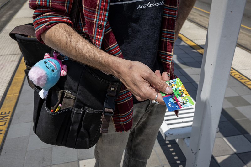 A transit rider holds a TAP card while standing on a Metro rail platform. A shoulder bag with pencils in an outer pocket and a small plush keychain hangs at their side.