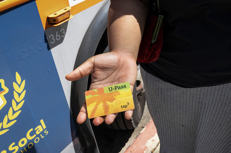 A hand holds a TAP U-Pass transit card beside the front wheel of a Metro bus, with part of a school district logo visible on the bus exterior.