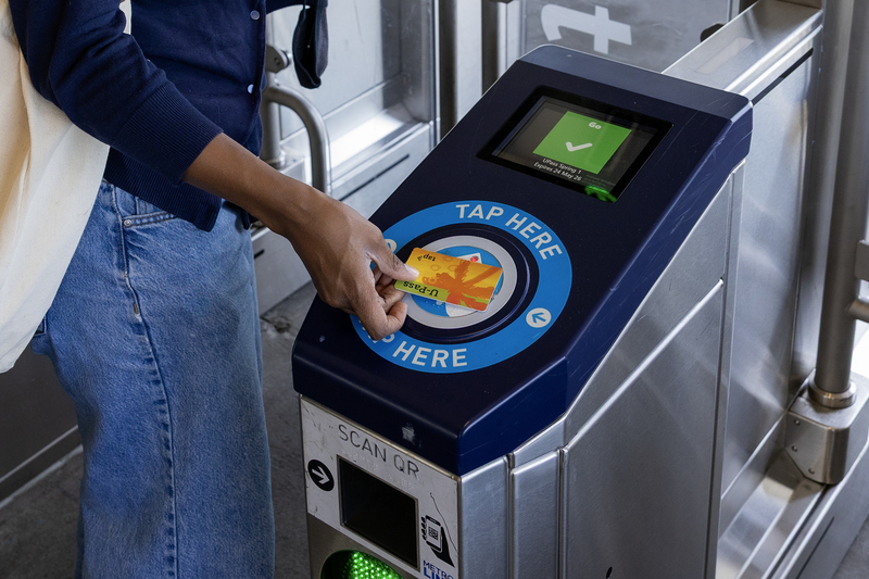 A transit rider taps a TAP U-Pass card on a fare reader at a Metro station turnstile, where the screen displays a green check mark confirming entry.