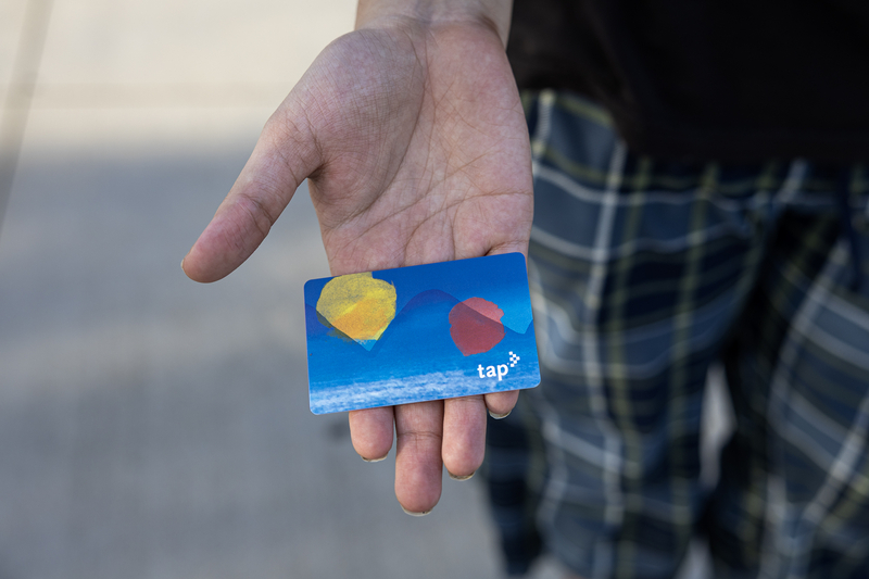 A hand holds a blue TAP transit card with an abstract design while standing outdoors, with only the rider's hand and plaid pants visible.