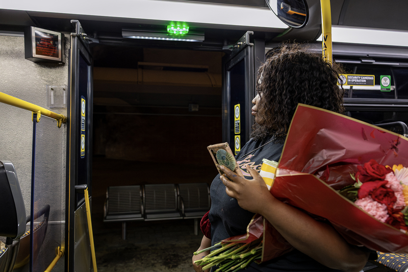 A transit rider stands near the open rear door of a Metro bus at night, holding a bouquet of flowers, a phone and a drink while looking out toward an empty bus stop.