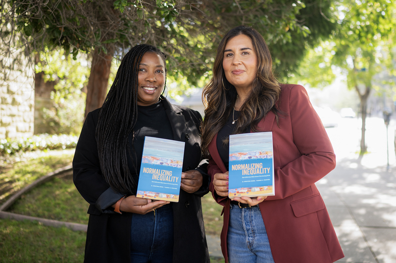 Two women stand on a tree-lined sidewalk holding copies of a book titled Normalizing Inequality. One wears a black jacket and the other a burgundy blazer, and both face the camera while posing with the book in front of a leafy residential streetscape.