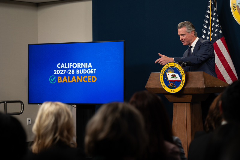 A suited public official speaks at a podium during a press conference, framed between blurred figures in the foreground. An American flag and part of the California state seal appear behind them as they gesture with their left hand while addressing the audience. A monitor with the words "California 2027-28 Budget Balanced" can be seen next to the public official.