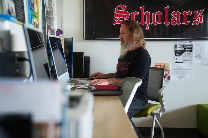A person with a long beard, wearing a black t-shirt that reads “Project Rebound” in red text, sits at a table, working on a computer in a row of computers set up against a wall inside a study room.