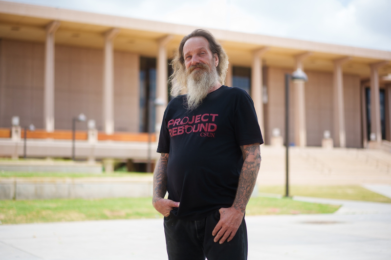 A person with a long beard, wearing a black t-shirt that reads “Project Rebound” in red text, stands with his hands halfway in his pockets in front of a college building, just out of focus in the background.