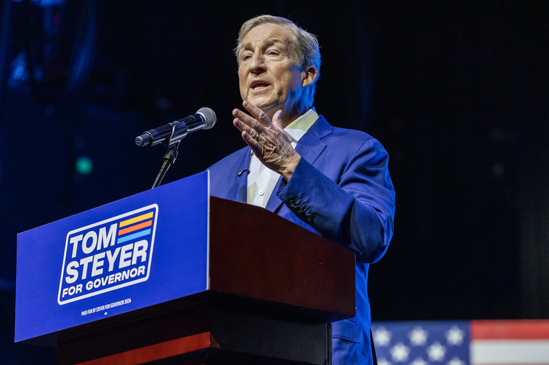 A person in a blue suit speaks at a podium with a “Tom Steyer for Governor” sign during a campaign event.